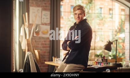 Ritratto di giovane uomo caucasico con capelli biondi e occhiali in posa per fotocamera in Creative Studio. Il talentuoso artista maschile attraversa le braccia e guarda la fotocamera con il sorriso. Concetto di occupazione moderna. Foto Stock