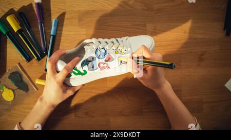 Foto con vista dall'alto: Un designer seduto dietro la scrivania di legno a casa e che applica immagini colorate sulle scarpe. Calzature eleganti fatte a mano e concetto di processo creativo. Primo piano. Foto Stock