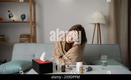 una giovane donna malata si siede sul divano di casa, si piega su un tavolo con delle pillole e tiene la testa con la mano. Concetto di freddo e mal di testa. Foto Stock