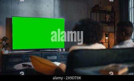Coppia afroamericana che guarda la TV con il Green Screen Mockup display. Sono seduti su un divano nel loro accogliente soggiorno e mangiano popcorn, godendo del servizio di film in streaming. Vista posteriore. Foto Stock