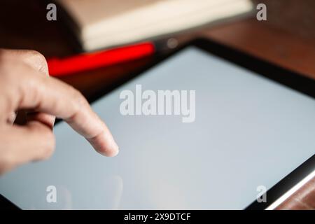 Primo piano di una mano utilizzando un tablet con schermo vuoto. Sullo sfondo sono presenti una penna rossa e un blocco appunti. Foto Stock