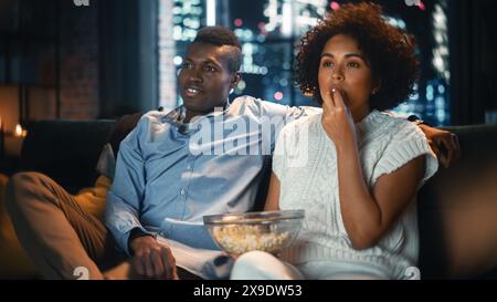Black Woman guarda il nuovo film e mangia popcorn a casa nell'elegante appartamento Loft. Il marito afroamericano è seduto accanto a lei sul divano. Concetto di intrattenimento online moderno. Foto Stock