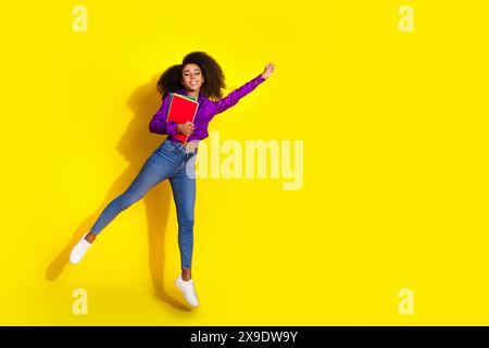 Foto a tutto il corpo di una giovane donna attraente che tiene libri che volano supereroina vestiti eleganti viola isolati su sfondo giallo Foto Stock