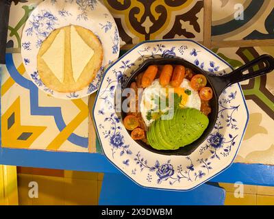 Vista dall'alto di una colazione tradizionale con uova fritte, fette di avocado, salsicce e pomodori ciliegini in una padella, servita con un piatto di che Foto Stock