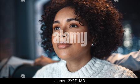 La bella giovane donna ride di notte e guarda Comedy Movie in TV. Il suo sfondo è sfocato. Ritratto di diverse donne multietniche che hanno un buon umore. Concetto di emozioni umane. Foto Stock