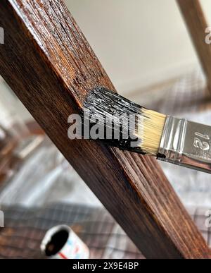 Primo piano di un pennello che applica vernice scura su una striscia di legno con molta consistenza. Sfondo sfocato e profondità di campo ridotta. Foto Stock