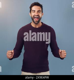 Ritratto, pugno e uomo in studio con festeggiamenti per il raggiungimento, il target o il raggiungimento dell'obiettivo con sfondo blu. Luce parassita, urla e uomo Foto Stock