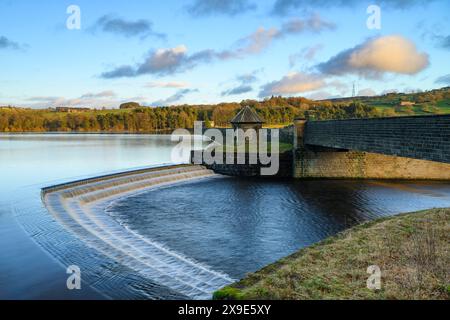 L'acqua scende giù per i gradini dal tranquillo e panoramico lago artificiale, sotto un ponte di pietra (soleggiato la sera, cielo blu e nuvole bianche) - Swinsty Reservoir, Inghilterra Regno Unito. Foto Stock