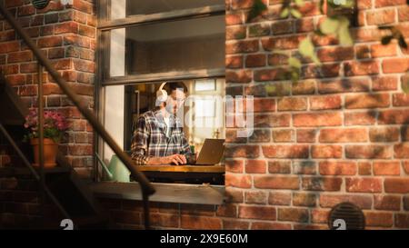 Moderna digitazione freelance su un notebook, ascolto di musica con le cuffie. Concetto di imprenditore e-Business professionale e di successo presso l'ufficio domestico. Vista dall'esterno verso la finestra. Foto Stock