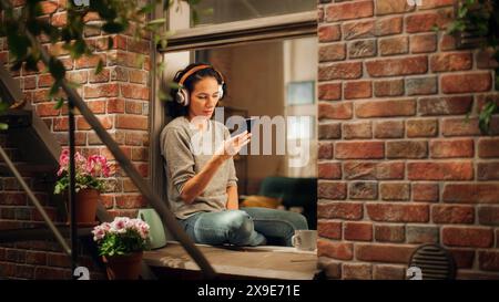 Bella donna brasiliana sorridente seduta su Windowsill e che utilizza le app per smartphone, navigare in Internet, cercare informazioni, servizio di incontri, sorridere felicemente. Attraverso la finestra dell'appartamento soleggiato. Foto Stock