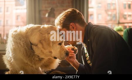 Giovane adulto di successo che gioca con il suo cane a casa, Active Golden Retriever. Uomo seduto su un pavimento che stuzzica, accarezza e gratta un cane eccitato, divertendosi nell'elegante soggiorno Loft. Foto Stock