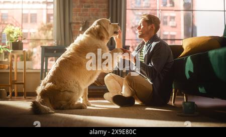 Happy Handsome Young Man offre al suo splendido Golden Retriever un regalo in cambio di un trucco o di un comando. Uomo attraente seduto su un pavimento che stuzzica, accarezza e gratta un cane eccitato. Foto Stock