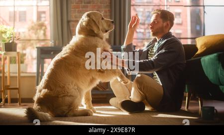 Happy Handsome Young Man offre al suo splendido Golden Retriever un regalo in cambio di un trucco o di un comando. Uomo attraente seduto su un pavimento che stuzzica, accarezza, gratta un cane eccitato. Foto Stock