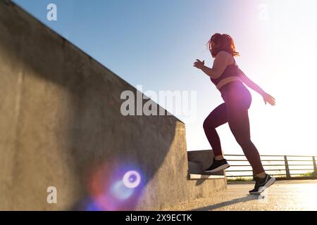Donna di taglia superiore che corre sulle scale durante un allenamento all'aperto con effetto flare dell'obiettivo, - determinazione e motivazione fitness, stile di vita sano, luminoso Foto Stock