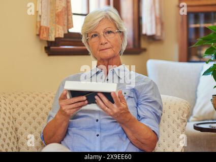 Incorniciatura, ritratto o donna anziana con foto a casa per ricordare, sperare o riposare sulla sedia del salotto. Rilassati, ricorda o solitaria anziana signora con Foto Stock