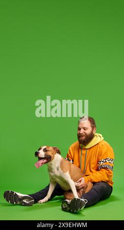 Poster. Uomo con cappuccio arancione seduto sul pavimento, coccolando il suo cane, che siede comodamente tra le braccia con la lingua fuori contro il vivace studio verde Foto Stock