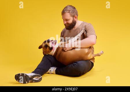 Giovane uomo che posa sdraiato sul pavimento e gioca con il suo delizioso e rilassato American Staffordshire Terrier sullo sfondo verde di uno studio. Foto Stock