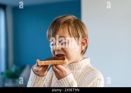 Un ragazzo morde in un panino con nocciole e cacao, seduto su un tavolo in salotto. Foto Stock