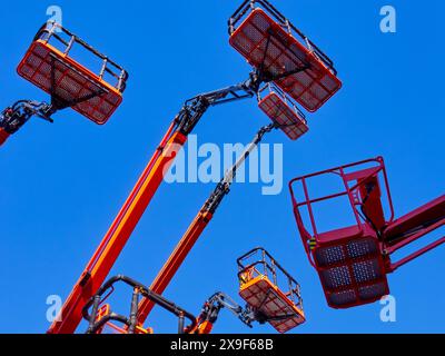 Gruppo numeroso di gru articolate, cestelli e bracci idraulici in alto in un cielo azzurro, con prospettiva ad angolo basso. Foto Stock