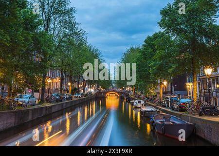 Una barca viaggia lungo un tipico canale nel cuore di Amsterdam, Paesi Bassi, il 27 maggio 2024 fiancheggiato da alberi, barche, auto parcheggiate come le luci begi Foto Stock