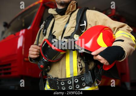 Vigile del fuoco in uniforme con casco e maschera vicino al camion dei pompieri rosso alla stazione, primo piano Foto Stock