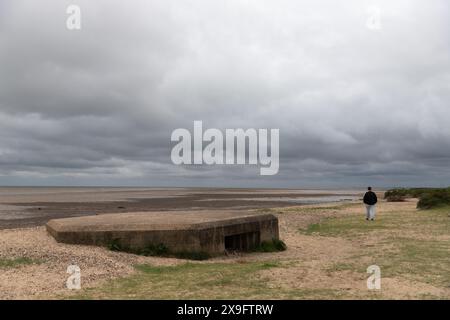 Una pillbox per mitragliatrici Vickers della seconda guerra mondiale, metà sepolta sulla spiaggia di Cudmore Grove, East Mersea, Mersea Island, Essex. Persona che cammina sullo sfondo Foto Stock