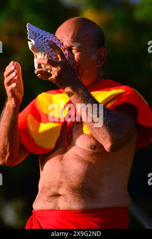 Un uomo anziano, vestito con un tradizionale abbigliamento hawaiano, segnala il tramonto soffiando in una conchiglia a conchiglia sulla spiaggia di Waikiki alle Hawaii Foto Stock