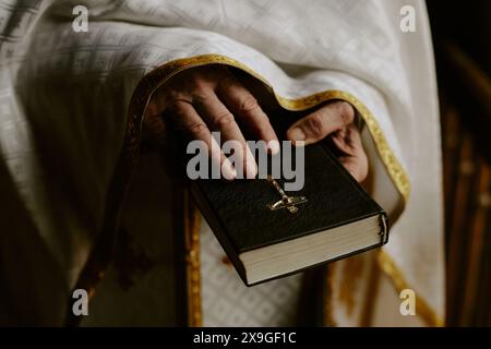 Primo piano delle mani rughe di un sacerdote cattolico anziano irriconoscibile che tiene il libro della Sacra Bibbia Foto Stock