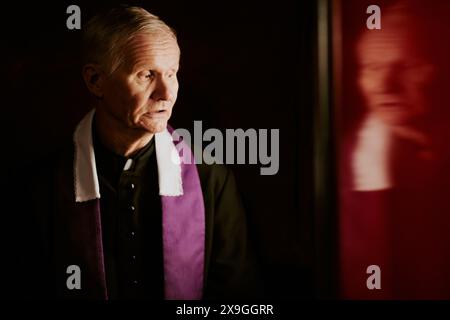 Primo piano medio di un anziano sacerdote cattolico caucasico che parla con il penitente in uno spazio confessionale, copia Foto Stock