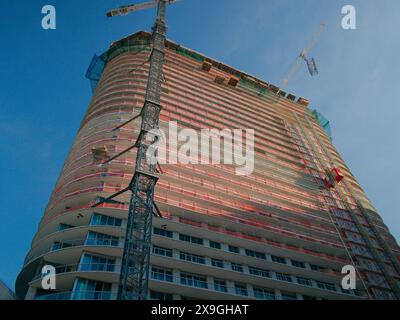 Ammira il lato dell'alto edificio a più piani e le gru in costruzione. Cielo blu e recinzione arancione. Niente persone e spazio per le copie. Gru temporanea Foto Stock