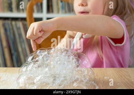 La bambina soffia bolle di schiuma di sapone, esplosioni, giochi divertenti per bambini, bolle di sapone fai da te, fare esperimenti, l'immaginazione del bambino accesa dalle meraviglie scien Foto Stock