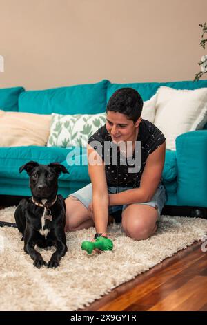 Vertical Una giovane donna giocosa con i capelli corti trascorre del tempo di qualità con il suo amato cane, partecipando a una divertente sessione di gioco sul pavimento accogliente della sua città Foto Stock