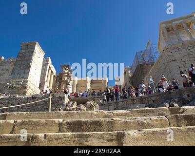 Molti turisti salgono i gradini di pietra di una rovina sull'Acropoli, antichi edifici con colonne e alberi sull'Acropoli di Atene contro a. Foto Stock