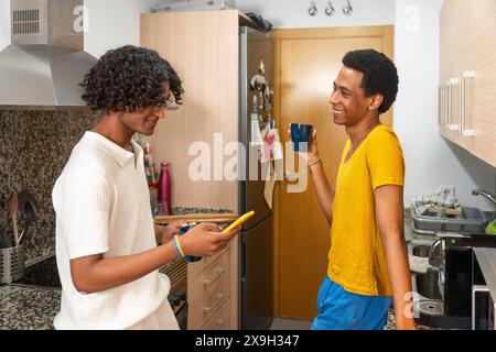 Scena domestica di una coppia gay che si gusta un caffè usando il telefono in cucina la mattina Foto Stock