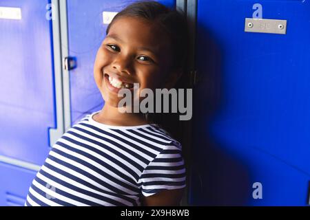 A scuola, giovane ragazza birazziale con un sorriso brillante in piedi accanto a armadietti blu Foto Stock