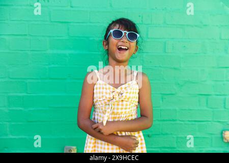 Una ragazza birazziale che indossa occhiali da sole e un abito da sole ride gioiosamente all'aperto Foto Stock