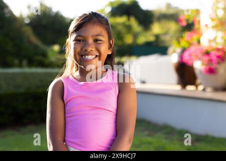 All'aperto, giovane ragazza birazziale sorride brillantemente mentre si trova in un giardino illuminato dal sole Foto Stock