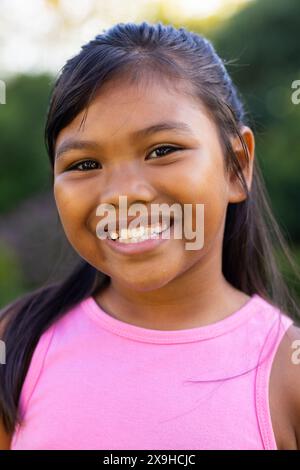 All'aperto, giovane ragazza birazziale che sorride brillantemente, indossa un top rosa con vegetazione Foto Stock