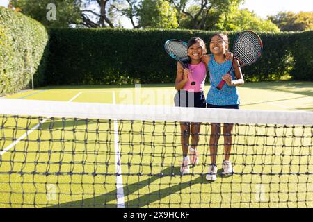 All'aperto, due giovani sorelle birazziali sorridenti, che tengono racchette da tennis sul campo da tennis Foto Stock