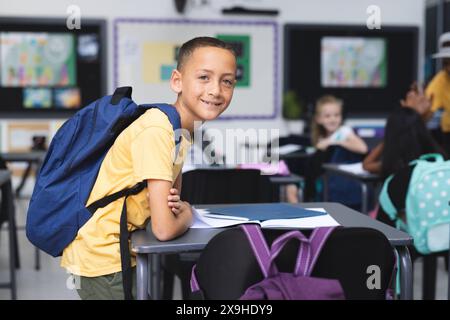 Un ragazzo birazziale con uno zaino sta in una classe scolastica, sorridente, con spazio per le copie. Circondato da compagni di classe, trasuda fiducia e prontezza Foto Stock