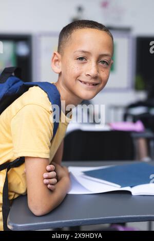 Un ragazzo birazziale con uno zaino si siede a una scrivania in un'aula scolastica, sorridendo Foto Stock