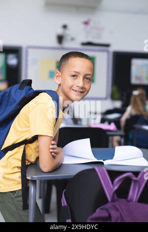 Un ragazzo birazziale con uno zaino sorride a una scrivania in un'aula scolastica Foto Stock