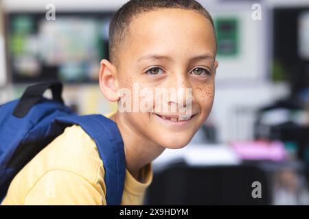 Ragazzo birazziale con sorrisi di lentiggini, indossa una camicia gialla e uno zaino in una classe scolastica Foto Stock