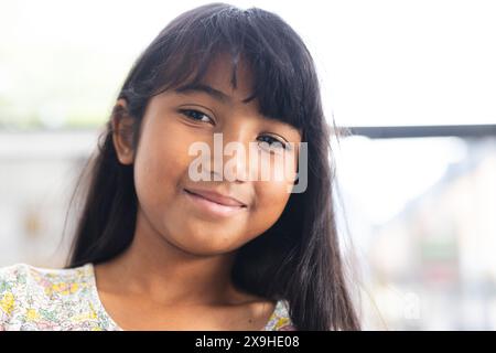 Ragazza birazziale con lunghi capelli marroni sorride dolcemente, indossando un abito floreale in una classe scolastica Foto Stock