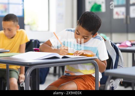Il ragazzo birazziale in una camicia a righe si concentra intensamente sulla scrittura in una classe scolastica Foto Stock