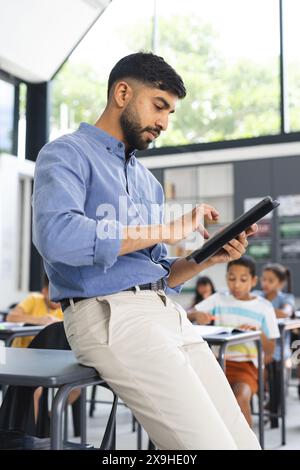 Il giovane insegnante asiatico con una camicia blu si concentra su un tablet in una classe scolastica Foto Stock