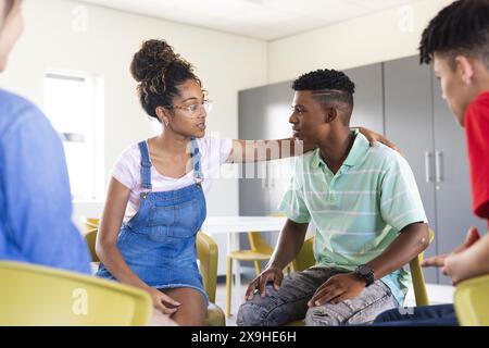 Un ragazzo birazziale adolescente conforta un altro in un ambiente di scuola superiore. Gruppi di studenti diversi si impegnano in una discussione di supporto al chiuso. Foto Stock