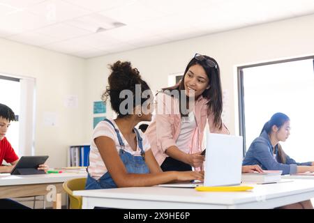 Una giovane insegnante birazziale insegna in classe, con spazio di copia. Si impegna con una ragazza adolescente indefinita e gli altri si concentrano sui loro studi. Foto Stock