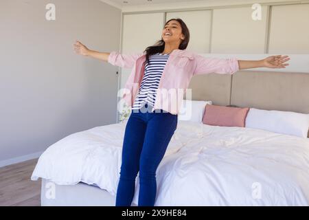 Una giovane donna birazziale a casa, in piedi sul suo letto, sorridente con le braccia tese Foto Stock