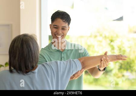 A casa, fisioterapista maschile birazziale e paziente anziano che fa esercizi al braccio. Camera luminosa e soleggiata con grandi finestre e vegetazione esterna Foto Stock
